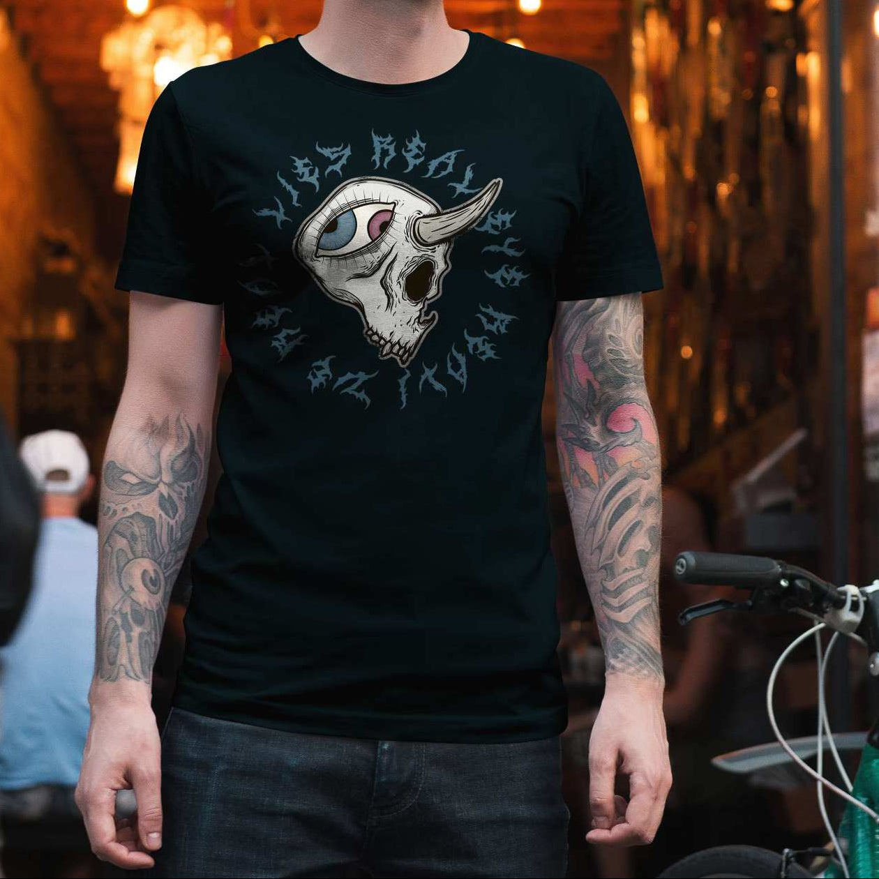 A person standing in a real-world urban alleyway wearing a black graphic t-shirt with a white skull and gothic lettering design, paired with dark jeans. 