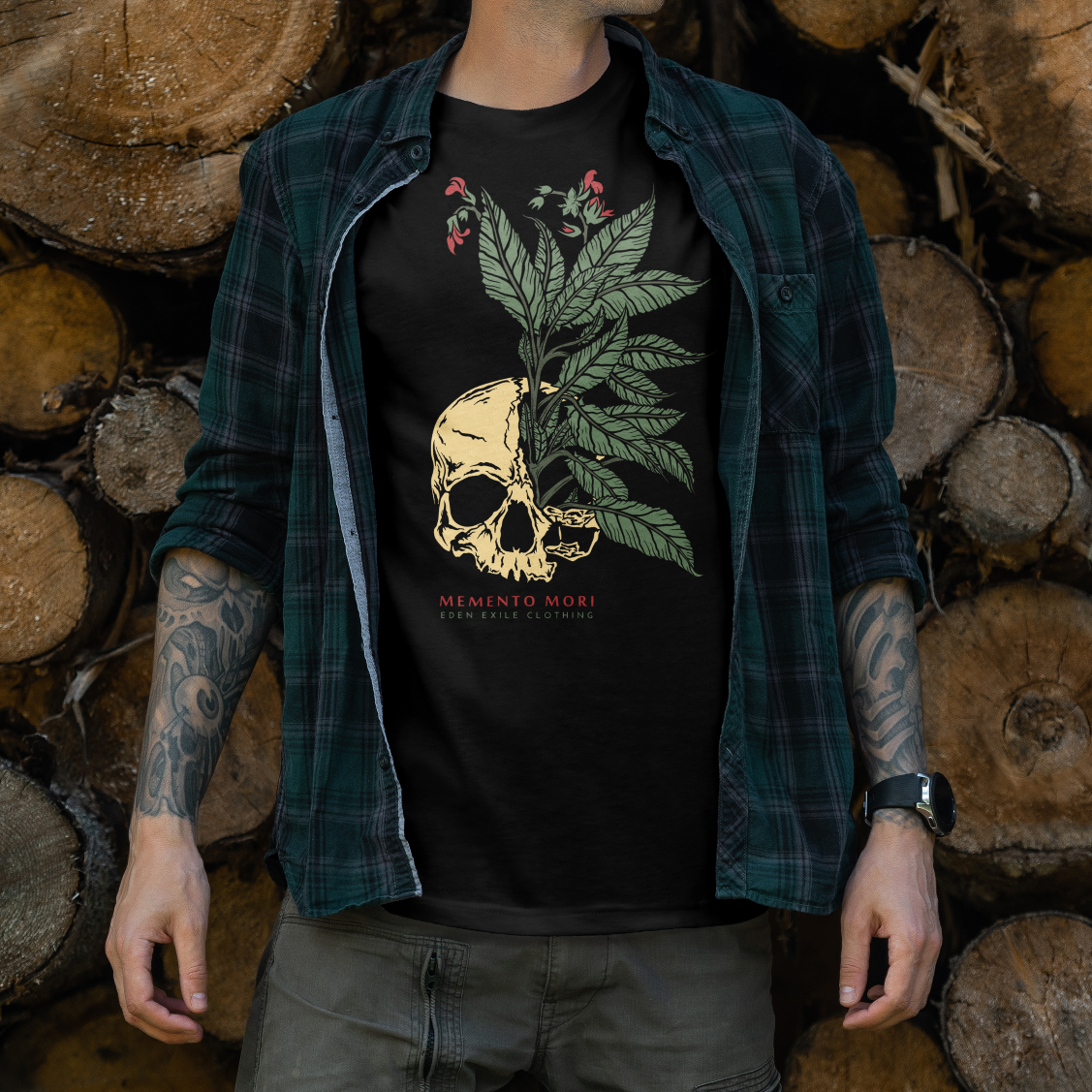 A man wearing a black graphic t-shirt with a skull and green leaf design, layered under an open dark green plaid flannel shirt, standing in front of a stacked pile of cut wood logs.