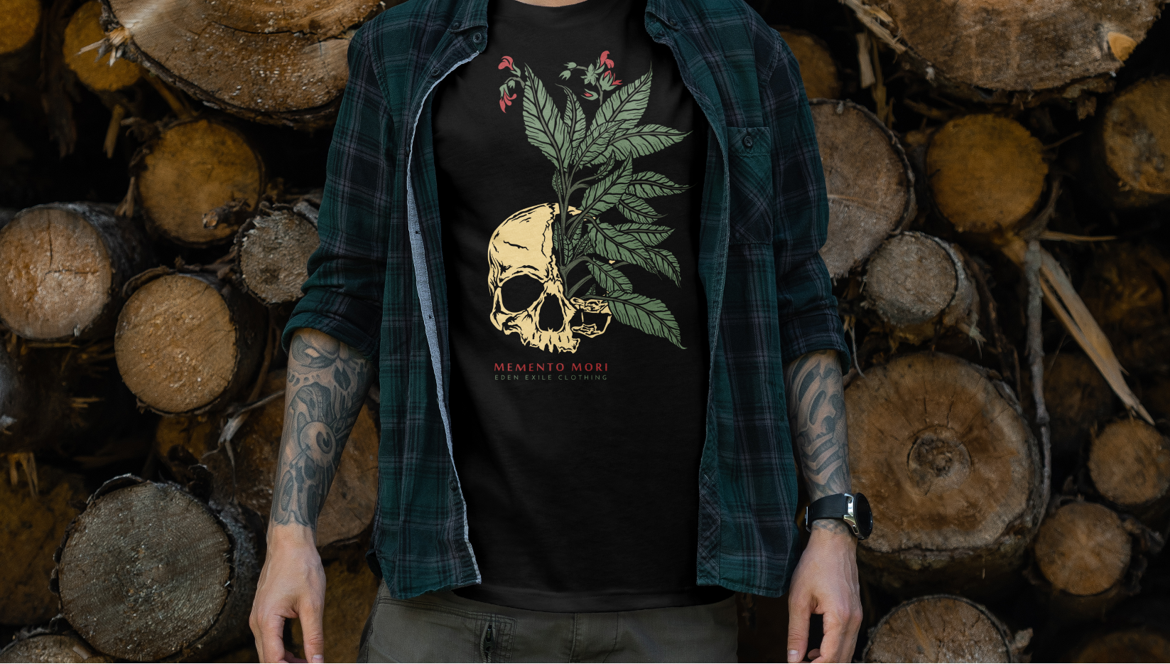 A man wearing a black graphic t-shirt with a skull and green leaf design, layered under an open dark green plaid flannel shirt, standing in front of a stacked pile of cut wood logs.