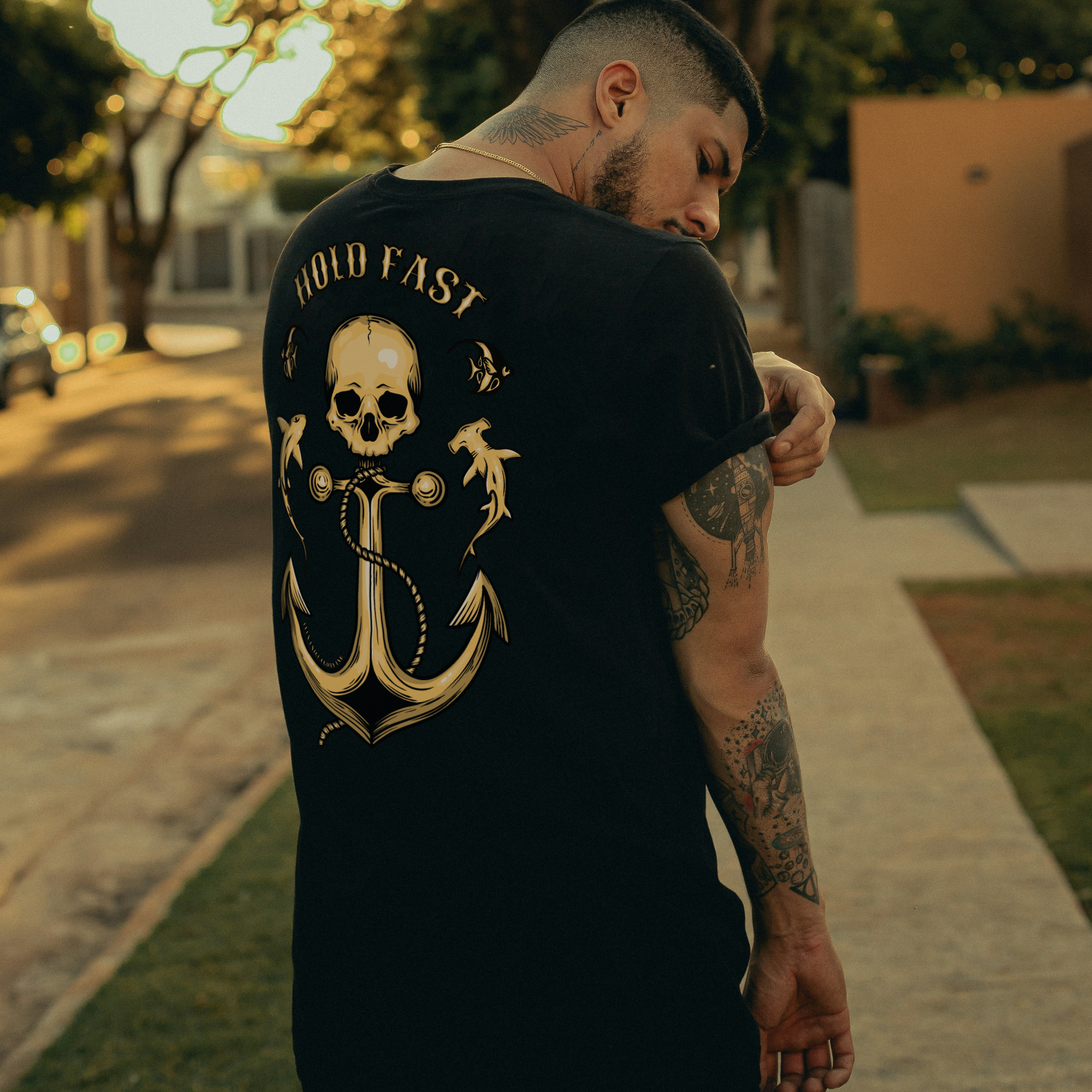 A man wearing a black t-shirt with a large gold anchor and skull graphic on the back, standing on a sidewalk in an outdoor setting with trees in the background.