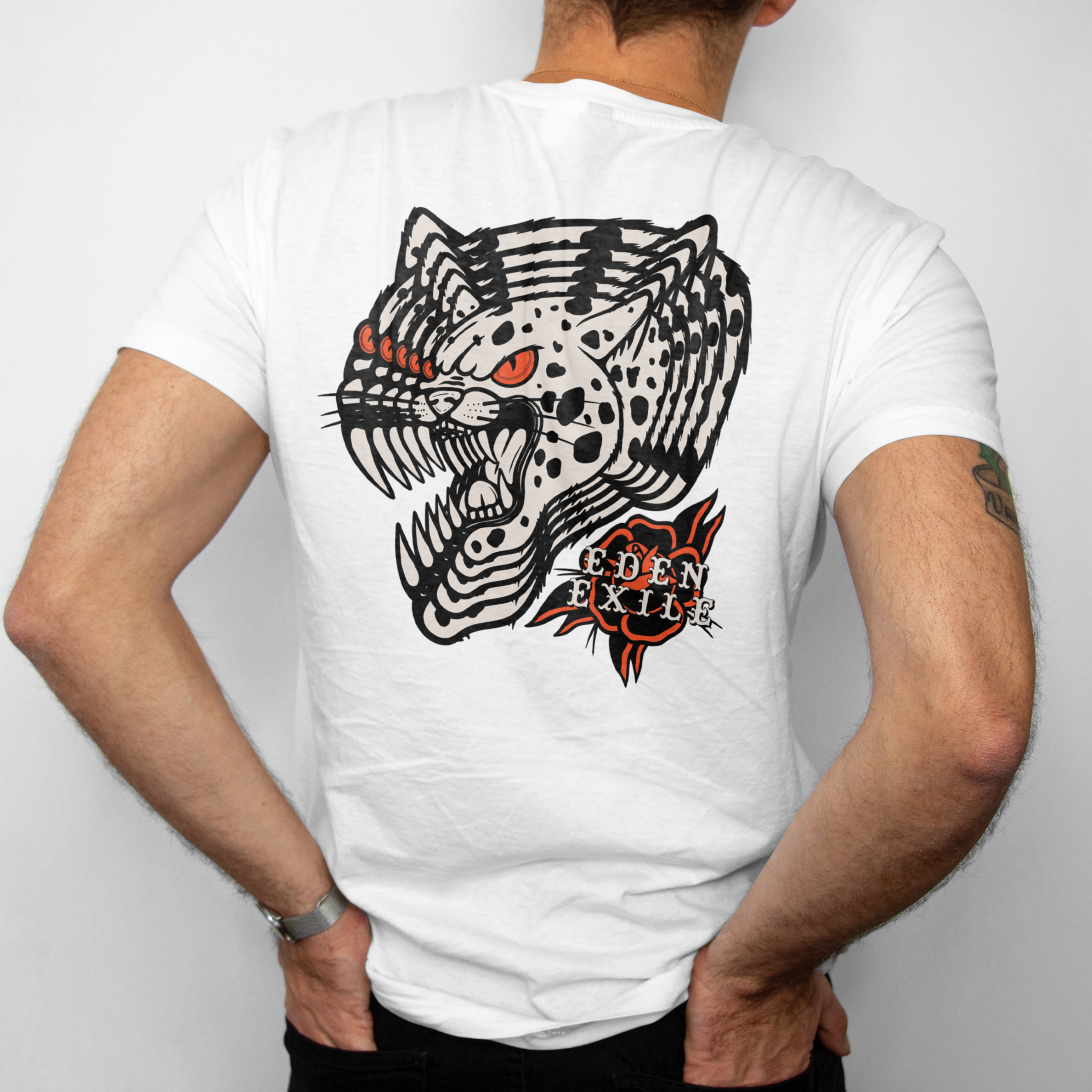 Back view of a man wearing a white t-shirt , showing a large graphic print of a black and white spotted big cat with red eyes and the text 'EDEN EXILE ' in a red and black Rose.