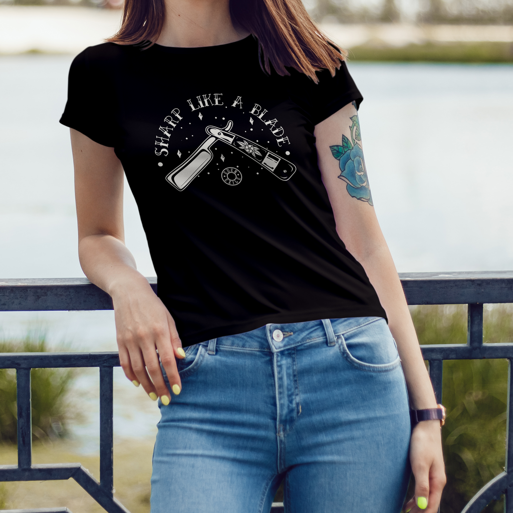 A woman wearing a black graphic t-shirt with a BNW vintage razor blade  design and the text 'sharp like a blade' paired with light blue jeans, standing outdoors by a railing with water in the background.