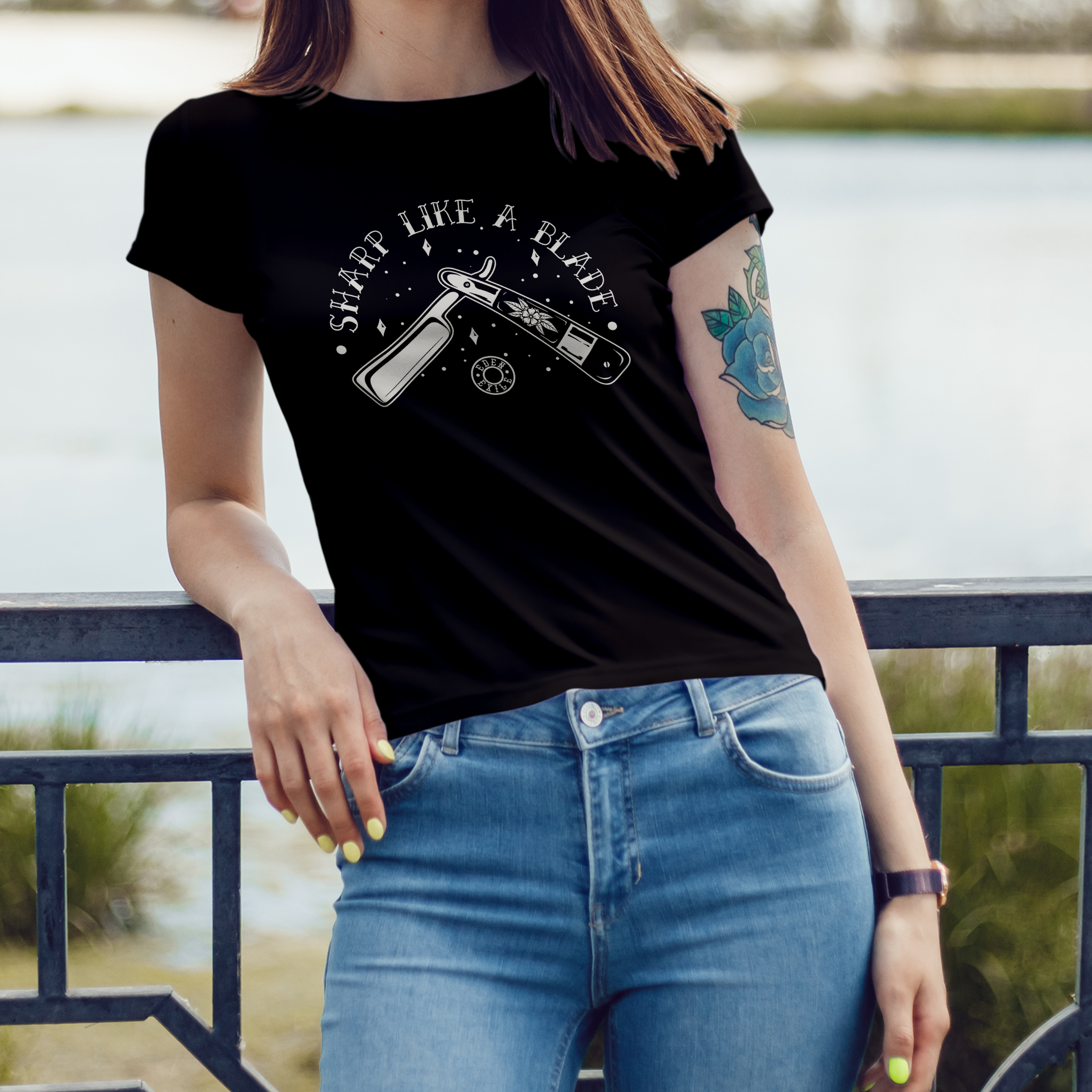A woman wearing a black graphic t-shirt with a BNW vintage razor blade  design and the text 'sharp like a blade' paired with light blue jeans, standing outdoors by a railing with water in the background.
