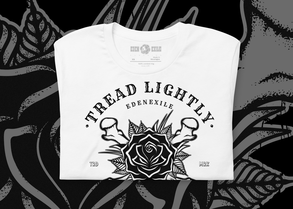 A folded white t-shirt with a black graphic print featuring a rose, mushrooms, and the text 'TREAD LIGHTLY' and 'EDENEXILE'. The shirt is displayed flat against a dark, high-contrast background