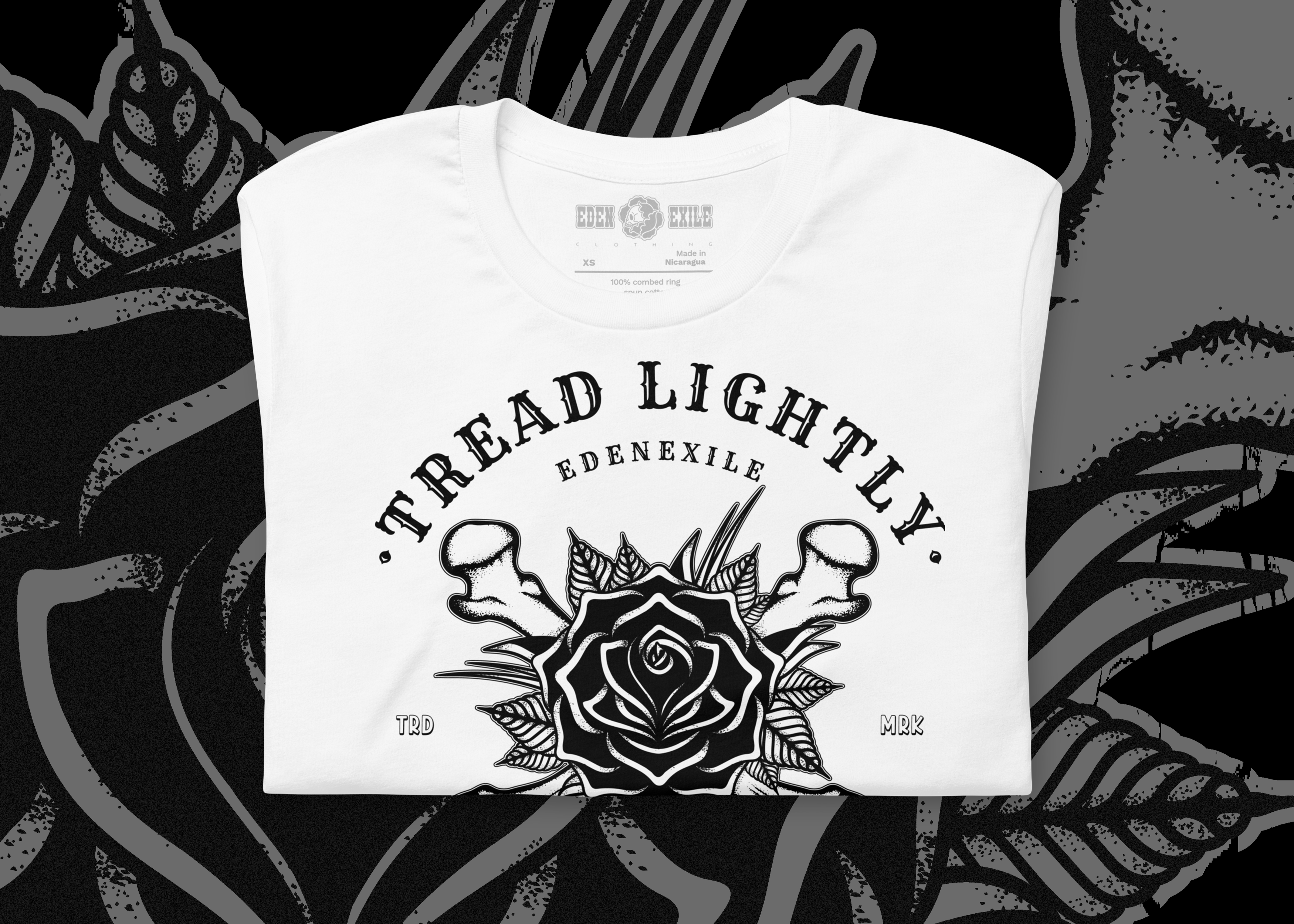 A folded white t-shirt with a black graphic print featuring a rose, mushrooms, and the text 'TREAD LIGHTLY' and 'EDENEXILE'. The shirt is displayed flat against a dark, high-contrast background