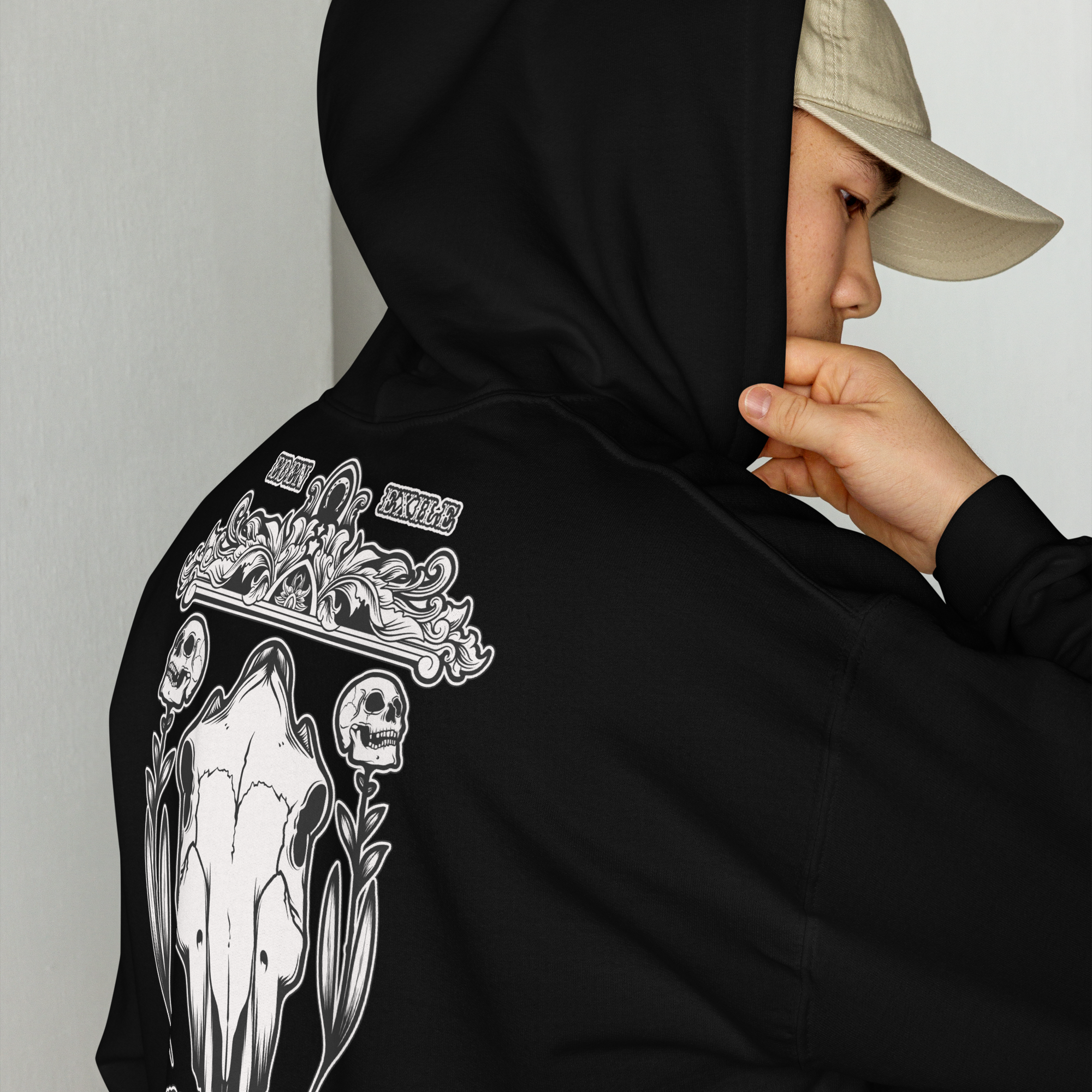 Person wearing a black hoodie with white design of horse skulls, renaissance wood carving art and "EDEN EXILE" text, looking away from the camera.