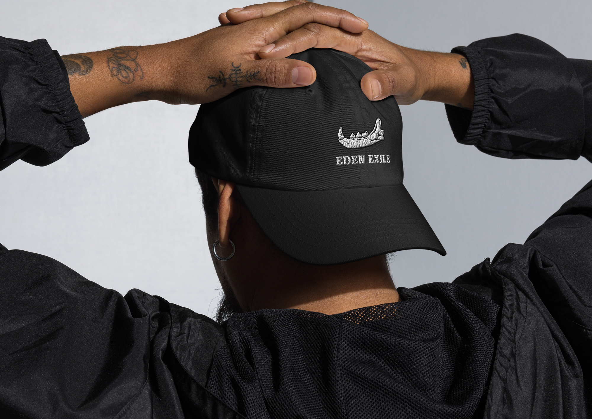 tattooed person wearing a black cap with an embroidered jaw bone design, inked hands clasped behind head