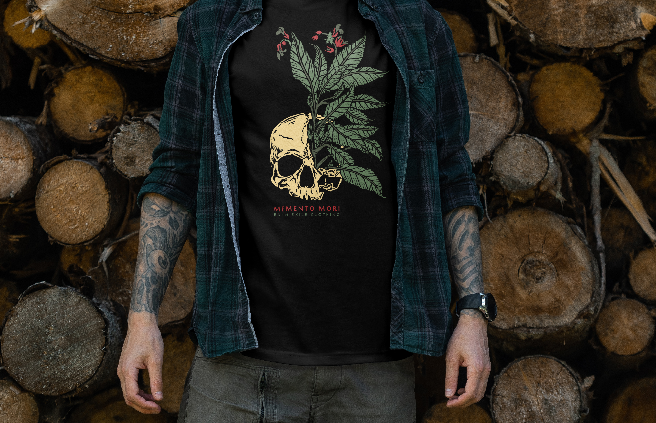 A man wearing a black graphic t-shirt with a skull and green leaf design , layered under an open dark green plaid flannel shirt, standing in front of a stacked pile of cut wood logs.